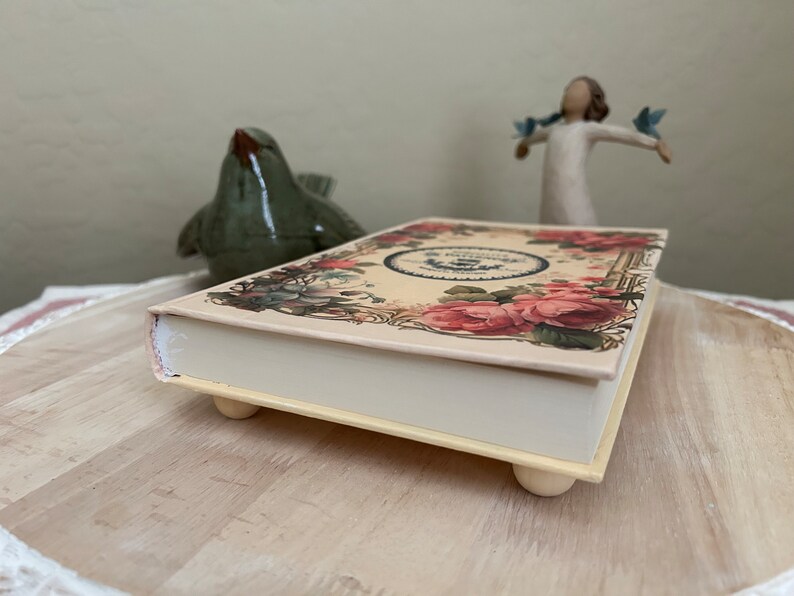 May include: A decorative box with a floral design and a white base. The box has four small feet and is made of wood. The box is closed and the top is decorated with a floral design.