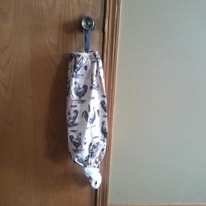 May include: A fabric bag with a drawstring closure, hanging from a door knob. The bag is cream-colored and patterned with black and gray chickens. The bag is designed to hold plastic grocery bags.