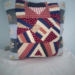 May include: A patchwork pillow with a heart-shaped design. The pillow features a mix of red, white, and blue fabrics, including plaid, stars, and stripes. Decorative buttons adorn the corners, adding a touch of detail to this handcrafted item.
