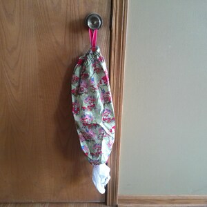 May include: A fabric bag with a floral pattern, hanging from a door knob. The bag is green with red, pink, and white floral designs. A pink ribbon is used to hang the bag. White tissue paper is visible at the bottom.