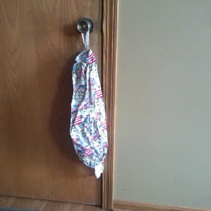 May include: A fabric bag with a patriotic print of red, white, and blue flags and stars, hanging from a silver door handle. The bag is gathered at the top and bottom, creating a cylindrical shape.