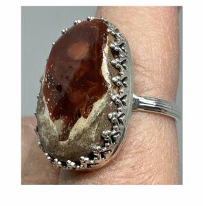 May include: An oval ring with a large, polished gemstone in shades of brown, beige, and cream. The stone is set in a silver-coloured metal band with a decorative floral border. The ring is displayed on a finger.
