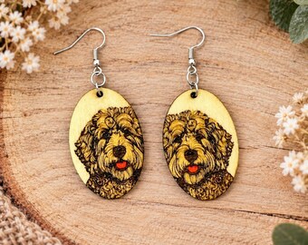 Puede incluir: Un par de pendientes ovalados de madera con una ilustración detallada grabada al fuego de un perro de pelo rizado con una lengua roja brillante. Los pendientes cuelgan de ganchos de metal plateado.