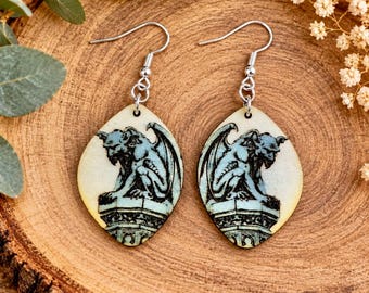 May include: A pair of teardrop-shaped earrings featuring a detailed illustration of a gargoyle perched on a stone pedestal. The earrings are crafted with a light-coloured base and silver-toned metal hooks for pierced ears.