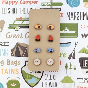 May include: A set of six wooden earrings with a camping theme. The earrings feature a tent, a campfire, a camper van, and a compass. The earrings are displayed on a brown card with the text "EATLUNE" and "CALL OF THE WILD".