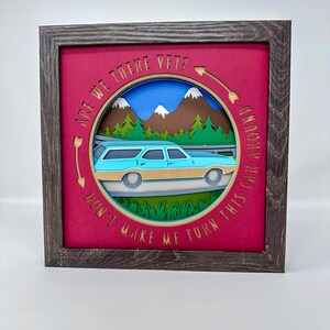 May include: A framed paper art piece with a light blue and tan station wagon, mountains, and trees. The artwork is layered and set against a magenta background. Text in a circle reads "ARE WE THERE YET? DON'T MAKE ME TURN THIS CAR AROUND."