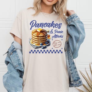 Könnte beinhalten: Ein beiges T-Shirt mit einer blauen Grafik eines Stapels Pfannkuchen mit Blaubeeren und Sirup. Der Text "Pancakes & Panic Attacks" befindet sich über den Pfannkuchen und "Buy one get one" unter den Pfannkuchen.