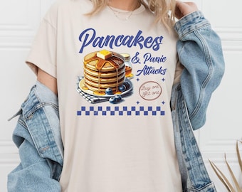 Puede incluir: Una camiseta beige con un gráfico azul de una pila de panqueques con arándanos y jarabe. El texto "Pancakes & Panic Attacks" está por encima de los panqueques y "Buy one get one" está por debajo de los panqueques.