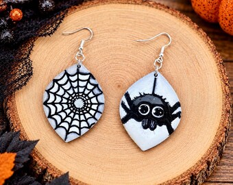 May include: A pair of dangle earrings featuring a leaf-shaped design. One earring displays a black spiderweb pattern on a white background, while the other shows a black spider with large white eyes on a white background, both with silver hooks.