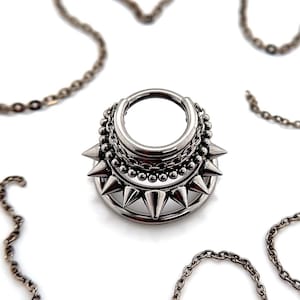 Puede incluir: Una pieza de joyería corporal plateada con un aro circular y un anillo exterior con púas. El aro está adornado con una cadena y detalles de cuentas.