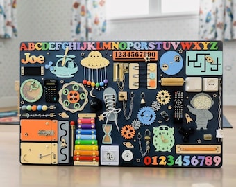 May include: A navy blue wooden activity board with colorful letters, numbers, and various interactive elements. Features a helicopter, a UFO, gears, a calculator, a clock, and other shapes. The name "Joe" is displayed in colorful letters.