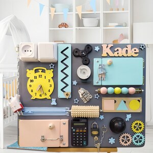 May include: A gray wooden activity board with various textures and activities for children. It features a clock, a zipper, a lock, a calculator, a spinning top, gears, a roller, and a nameplate that says "Kade".