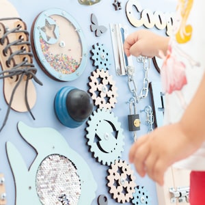 May include: A blue and beige wooden activity board with various textures and activities for children. The board features a shoe with laces, a spinning gear, a lock and chain, a spinning knob, and a small window with beads inside. The board is labeled "Coop" in white letters.