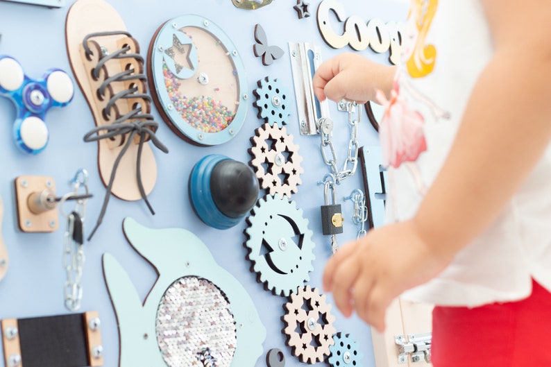 May include: A blue and beige wooden activity board with various textures and activities for children. The board features a shoe with laces, a spinning gear, a lock and chain, a spinning knob, and a small window with beads inside. The board is labeled "Coop" in white letters.