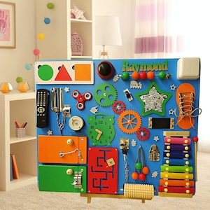 May include: A blue wooden activity board with various textures and activities for children. It features a clock, a chain, a lock, a maze, a xylophone, a spinner, a gear, a star, a shoe, a light switch, a remote control, and a knob. The board is personalized with the name "Raymond".