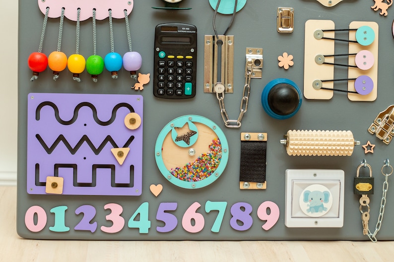 May include: A colorful wooden activity board for toddlers with a variety of textures and activities, including a calculator, a chain, a rolling pin, a lock, a maze, a star-shaped spinner, buttons, and numbers 0-9.