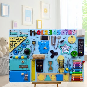 May include: A blue and yellow wooden busy board with various activities for children, including a clock, a lock, a chain, a star, a xylophone, a calculator, a zipper, a door latch, a maze, a roller, a spinner, and a set of numbers. The board is labeled "Rain River".