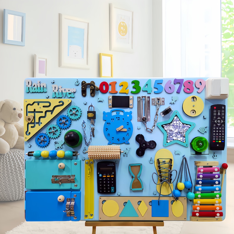 May include: A blue and yellow wooden busy board with various activities for children, including a clock, a lock, a chain, a star, a xylophone, a calculator, a zipper, a door latch, a maze, a roller, a spinner, and a set of numbers. The board is labeled "Rain River".