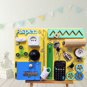 May include: A yellow wooden busy board with various activities for children, including a light switch, an electrical outlet, a zipper, a chain, a calculator, a lock, a knob, a spinner, a roller, gears, and a name tag that says "Aspen".