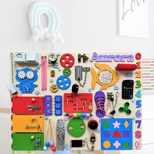May include: A colorful wooden activity board with various textures and activities for children. It features a clock, gears, a zipper, a lock, a chain, a maze, a fish, a calculator, a fidget spinner, a lacing shoe, a light switch, a door knob, and a remote control. The board is personalized with the name "Aurora" in purple letters.