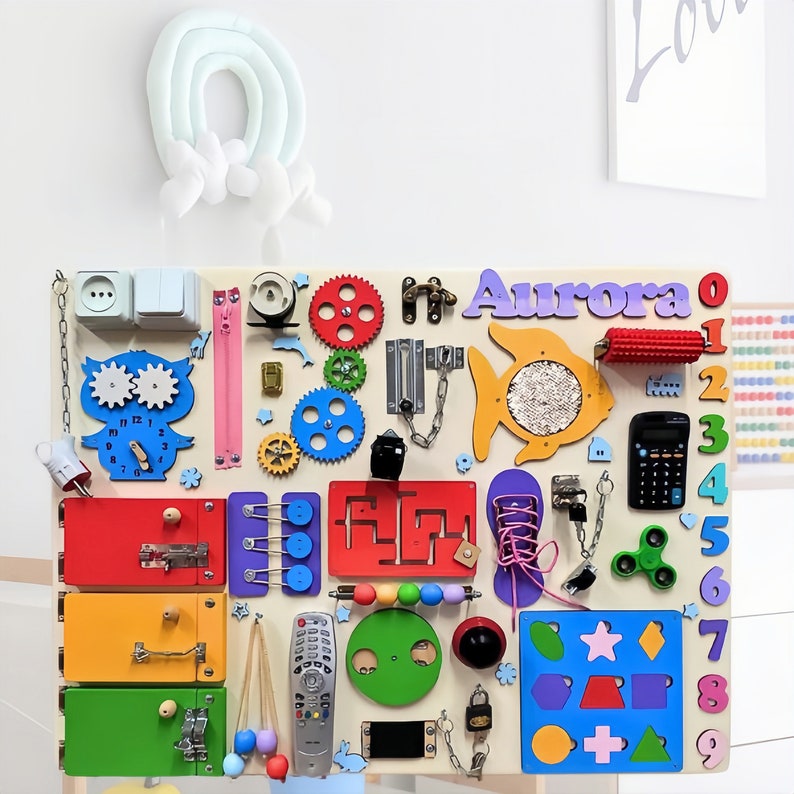 May include: A colorful wooden activity board with various textures and activities for children. It features a clock, gears, a zipper, a lock, a chain, a maze, a fish, a calculator, a fidget spinner, a lacing shoe, a light switch, a door knob, and a remote control. The board is personalized with the name "Aurora" in purple letters.