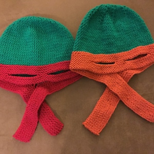 May include: Two knitted hats in the style of the Teenage Mutant Ninja Turtles. One hat is green and red, the other is green and orange. Each hat has eye slits and long ties. The hats are made of a textured knit fabric.