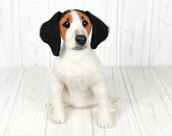 May include: A white, black, and brown needle felted dog figurine. The dog is sitting with its front paws in front of it and its head tilted slightly to the side. The dog has black ears and a black nose.
