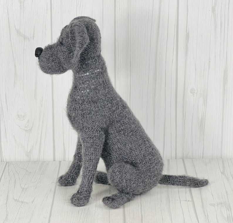Puede incluir: Una figurita de perro de punto gris sentada sobre una superficie de madera blanca. El perro est&aacute; en posici&oacute;n sentada con la cola enroscada alrededor de sus patas.