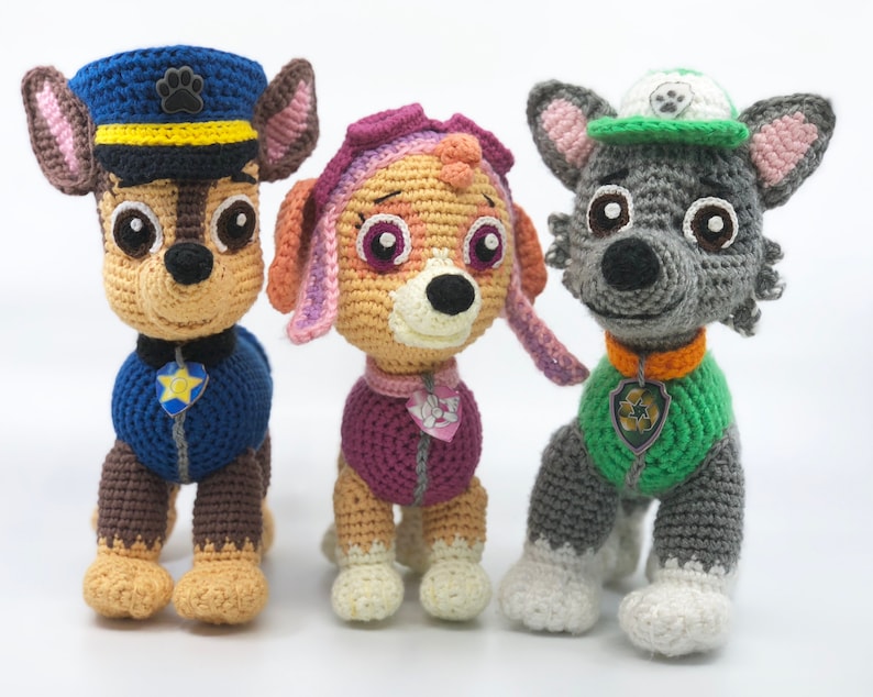 Op de afbeelding: Drie gehaakte knuffels, een bruine en beige hond met een blauwe hoed en uniform, een roze en beige hond met een paarse hoed en jas, en een grijze wolf met een groene hoed en jas.