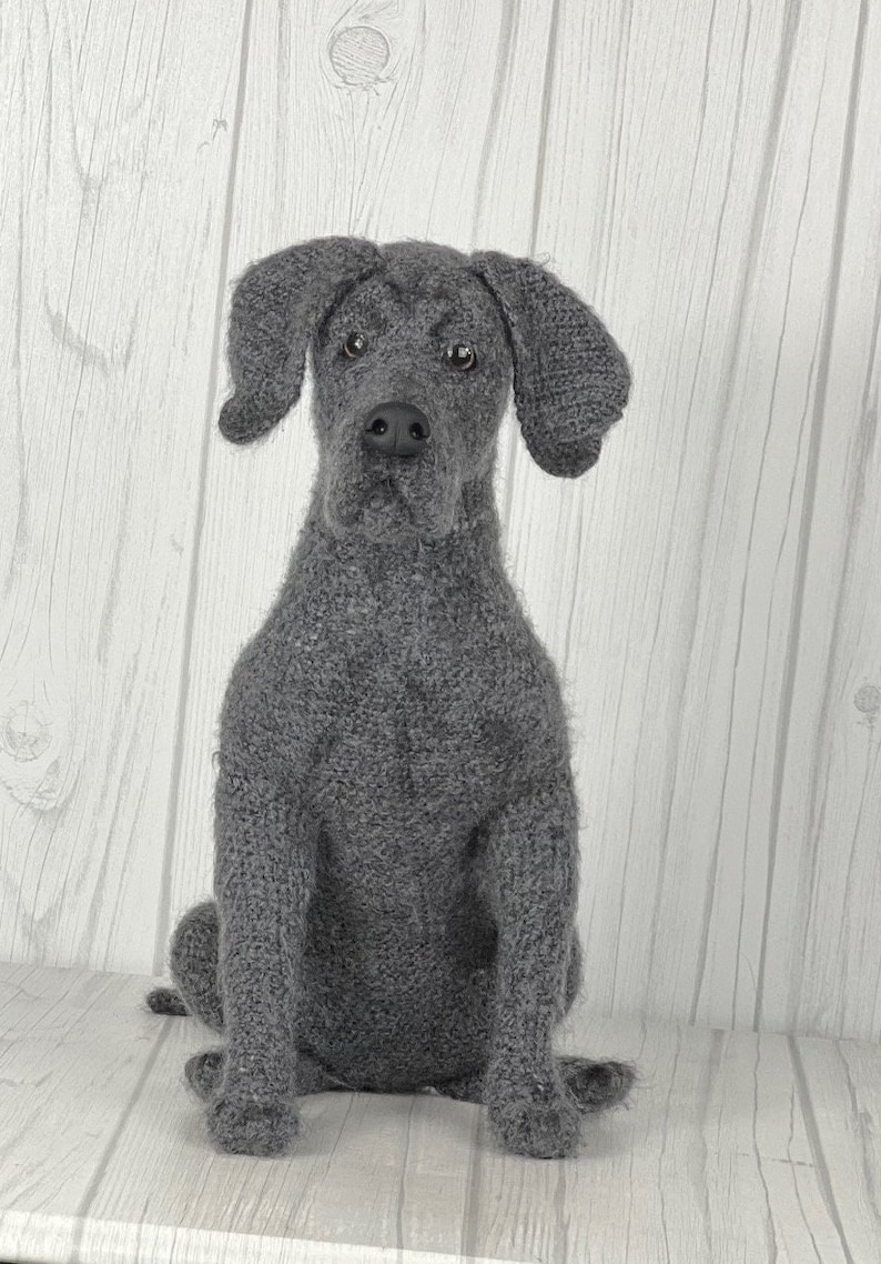 Puede incluir: Un juguete de perro de punto gris en forma de Weimaraner. El juguete de perro est&aacute; sentado sobre una superficie de madera blanca.
