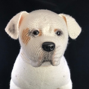 May include: A crocheted white dog with brown and black markings on its face. The dog has a black nose and is looking directly at the camera.
