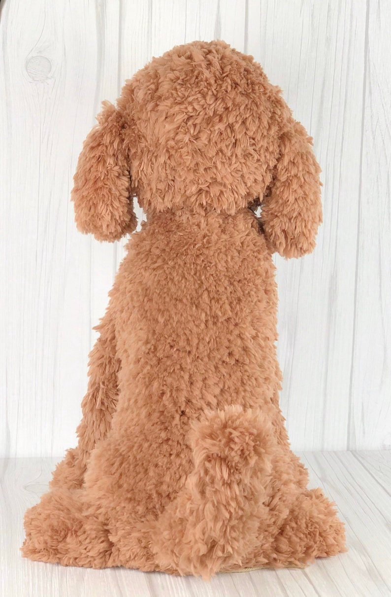 以下が含まれることがあります： 白い木製の表面に座っている茶色のぬいぐるみ犬。犬はふわふわの縮れた毛皮と長い尻尾をしています。