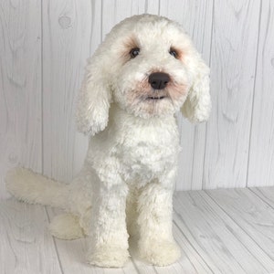 以下が含まれることがあります： 茶色の目と鼻を持つ、白いふわふわのぬいぐるみ犬。犬は尻尾を後ろに巻いて座っている。