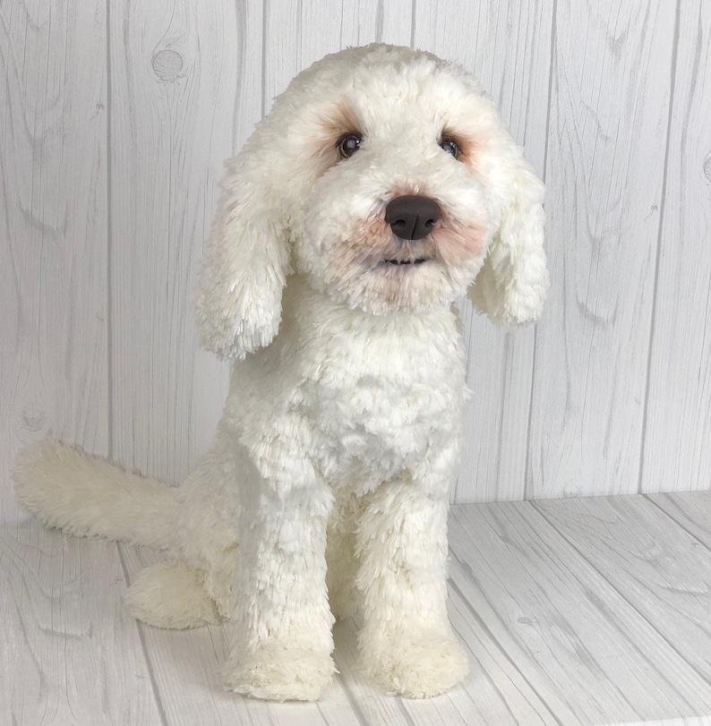 以下が含まれることがあります： 茶色の目と鼻を持つ、白いふわふわのぬいぐるみ犬。犬は尻尾を後ろに巻いて座っている。