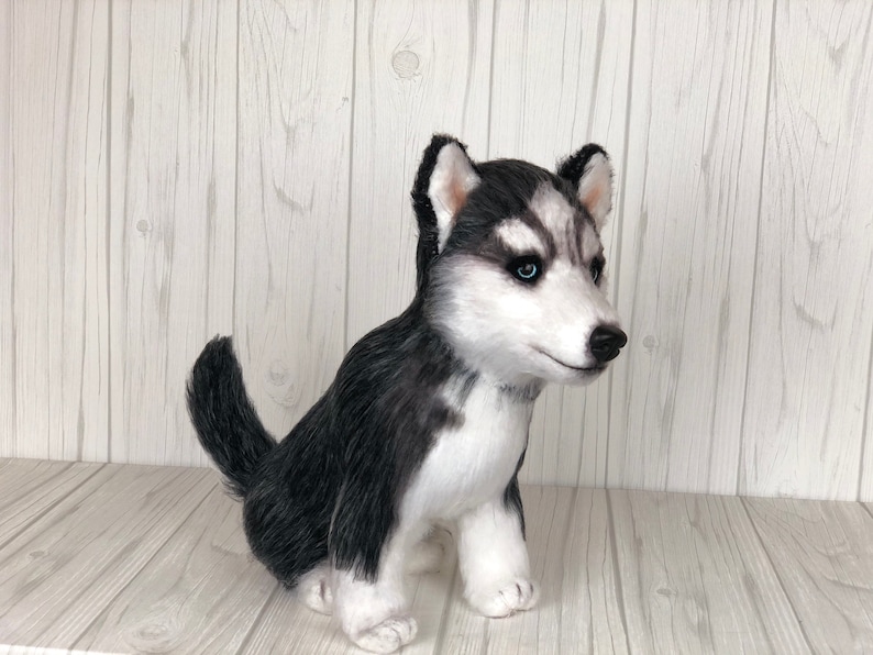 May include: A black and white stuffed husky puppy sitting on a white wooden surface. The puppy has blue eyes and a black nose.