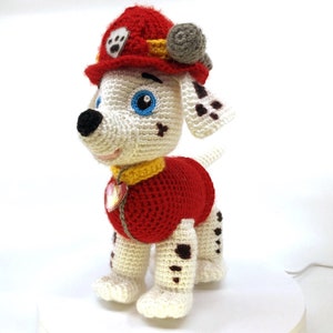 Op de afbeelding: Een wit-zwart dalmati&euml;r knuffeldier met een rood en geel brandweerpak met een blauw oog en een rode muts.