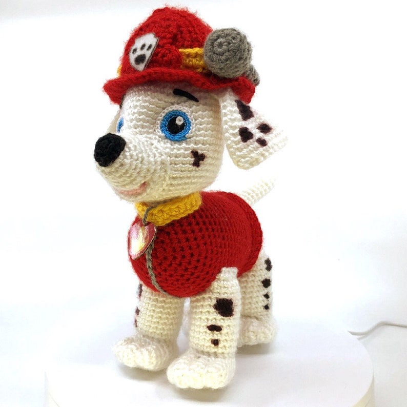 Op de afbeelding: Een wit-zwart dalmati&euml;r knuffeldier met een rood en geel brandweerpak met een blauw oog en een rode muts.