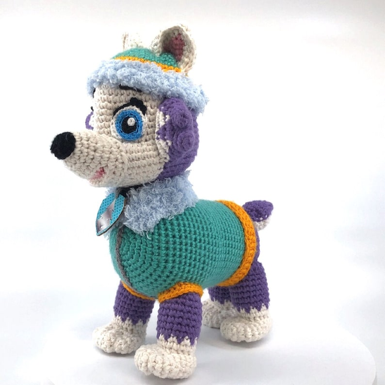 Op de afbeelding: Een gehaakt knuffelhondje met een wit, blauw en paars outfit. De hond heeft een blauw oog en een zwarte neus.