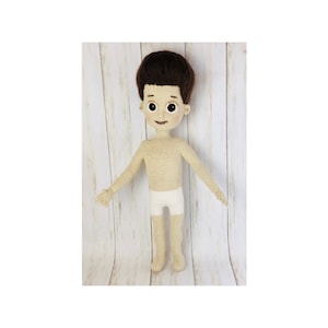 May include: A crocheted doll with brown hair, a beige body, and white underwear. The doll is standing on a white wooden surface.