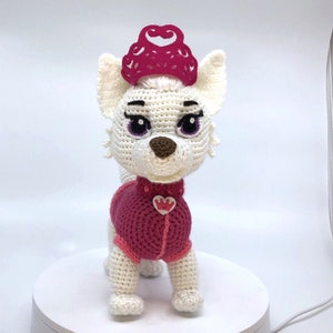 May include: White crocheted plush toy dog wearing a pink crocheted dress and a pink crown. The dog has purple eyes and a brown nose.