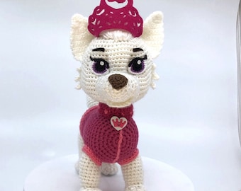 Può includere: Un cane di peluche bianco all'uncinetto che indossa un vestito rosa all'uncinetto e una corona rosa. Il cane ha gli occhi viola e un naso marrone.