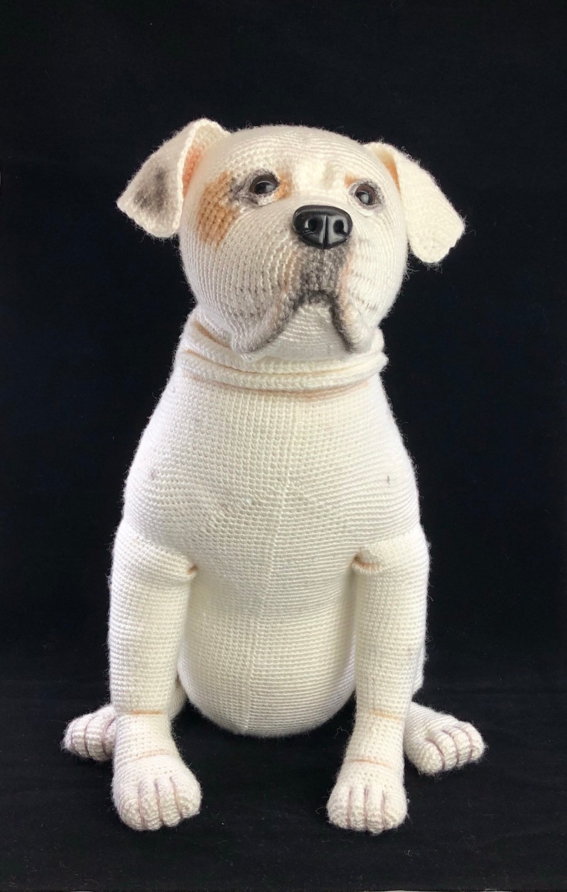May include: White crocheted stuffed dog sitting with its paws crossed. The dog has brown and black markings on its face.