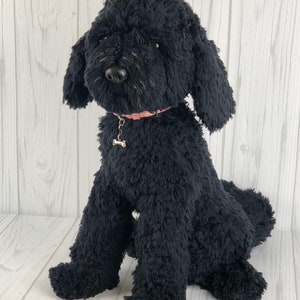 以下が含まれることがあります： 白い木製の背景に、黒いぬいぐるみプードル犬が座っています。犬は、骨のチャームが付いたピンクの首輪をしています。