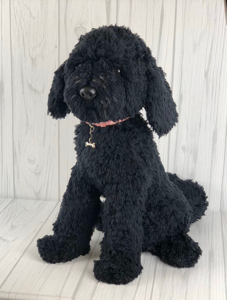 以下が含まれることがあります： 白い木製の背景に、黒いぬいぐるみプードル犬が座っています。犬は、骨のチャームが付いたピンクの首輪をしています。