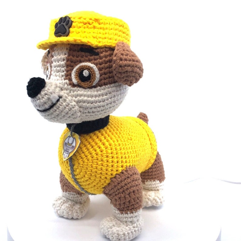 Op de afbeelding: Een gehaakte knuffelhond met een gele bouwvakkershoed en outfit. De hond heeft bruin en wit haar en een zwarte halsband met een geel label.