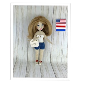 May include: A crocheted doll with blonde hair, wearing a white top, blue denim shorts, and red shoes. The doll is holding a white crocheted bag. The doll is standing on a wooden surface. There are two flags in the background, an American flag and a Dutch flag.