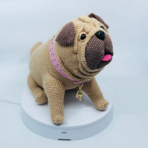 Puede incluir: Un juguete de perro pug de crochet con un collar rosa y un dije dorado. El perro está sentado en una plataforma blanca.