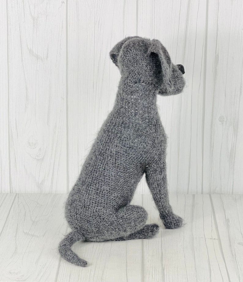 Puede incluir: Un juguete de perro de punto gris en posici&oacute;n sentada. El juguete est&aacute; hecho de un material suave y esponjoso y tiene una cola larga.