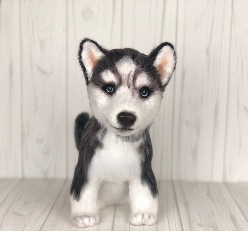 May include: A plush toy husky puppy with black and white fur and blue eyes. The puppy is standing on a white wooden surface.