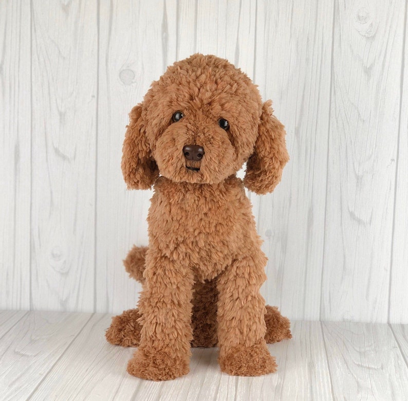 以下が含まれることがあります： 茶色のふわふわしたプードルのぬいぐるみ。白い木製の表面に座っています。